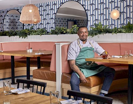 A person in a green apron sits on a booth edge in a stylish restaurant with patterned walls, plants, and modern decor.