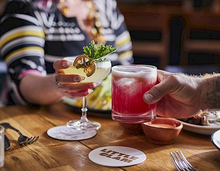 Two people clinking glasses with cocktails over a table. Plates of food and a coaster saying 'Little Giant' are visible.