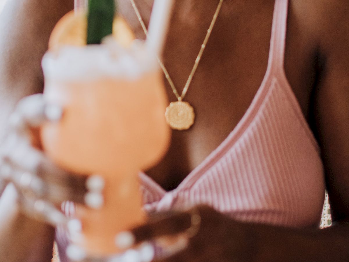 A person in a pink top holds a cocktail garnished with a slice of fruit and a leaf, using a straw.