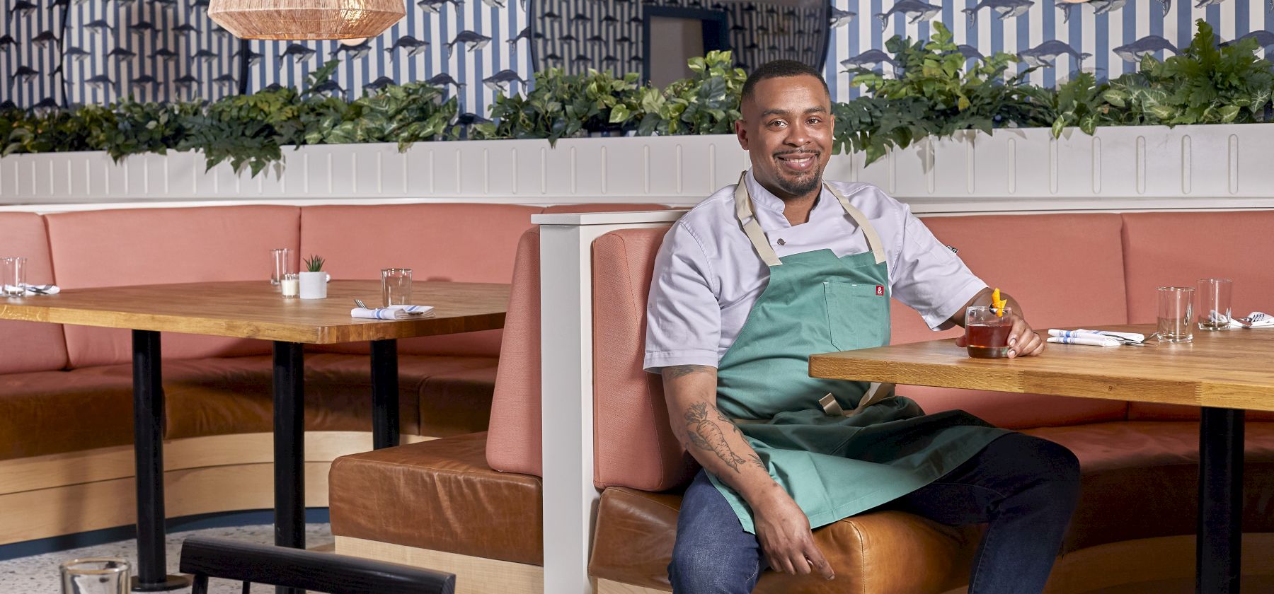 A person in a green apron sits on a restaurant booth, with patterned walls, hanging lamp, and greenery in the background.