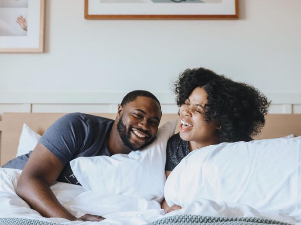 A couple is smiling and relaxing in bed, with framed art pieces on the wall behind them.