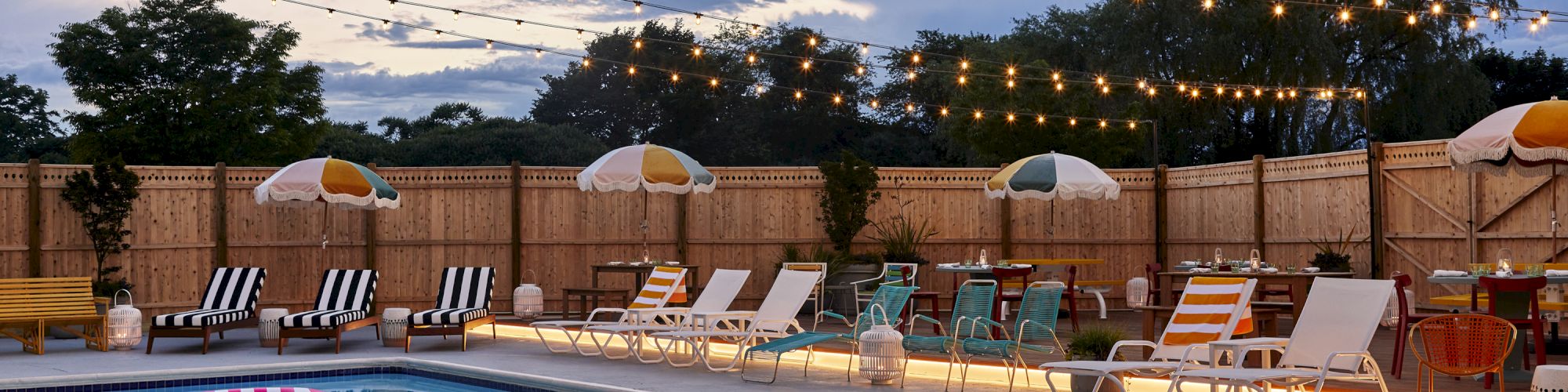 A swimming pool with lounge chairs, tables, and string lights against a sunset sky, featuring umbrellas.