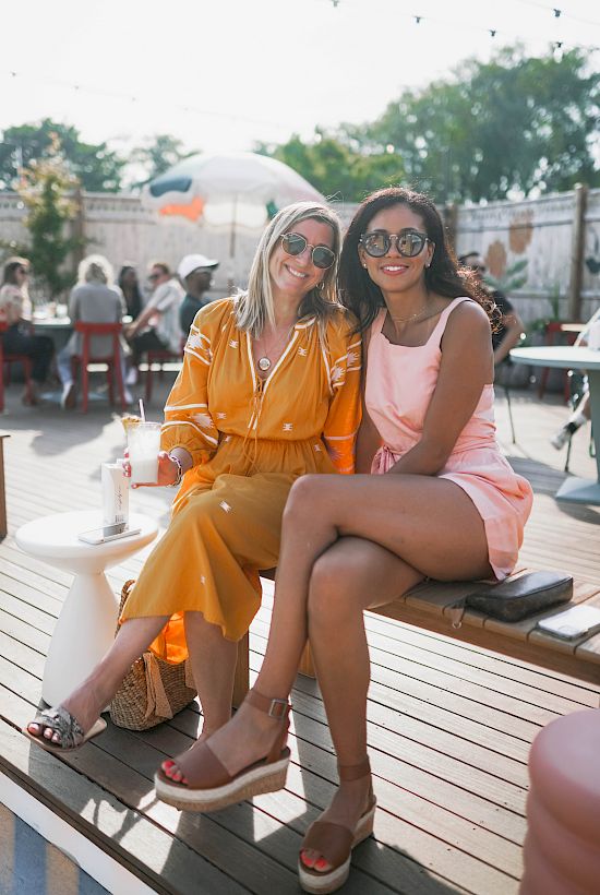 Two people sitting on an outdoor bench, wearing sunglasses and summer clothes, surrounded by other people and trees in the background.