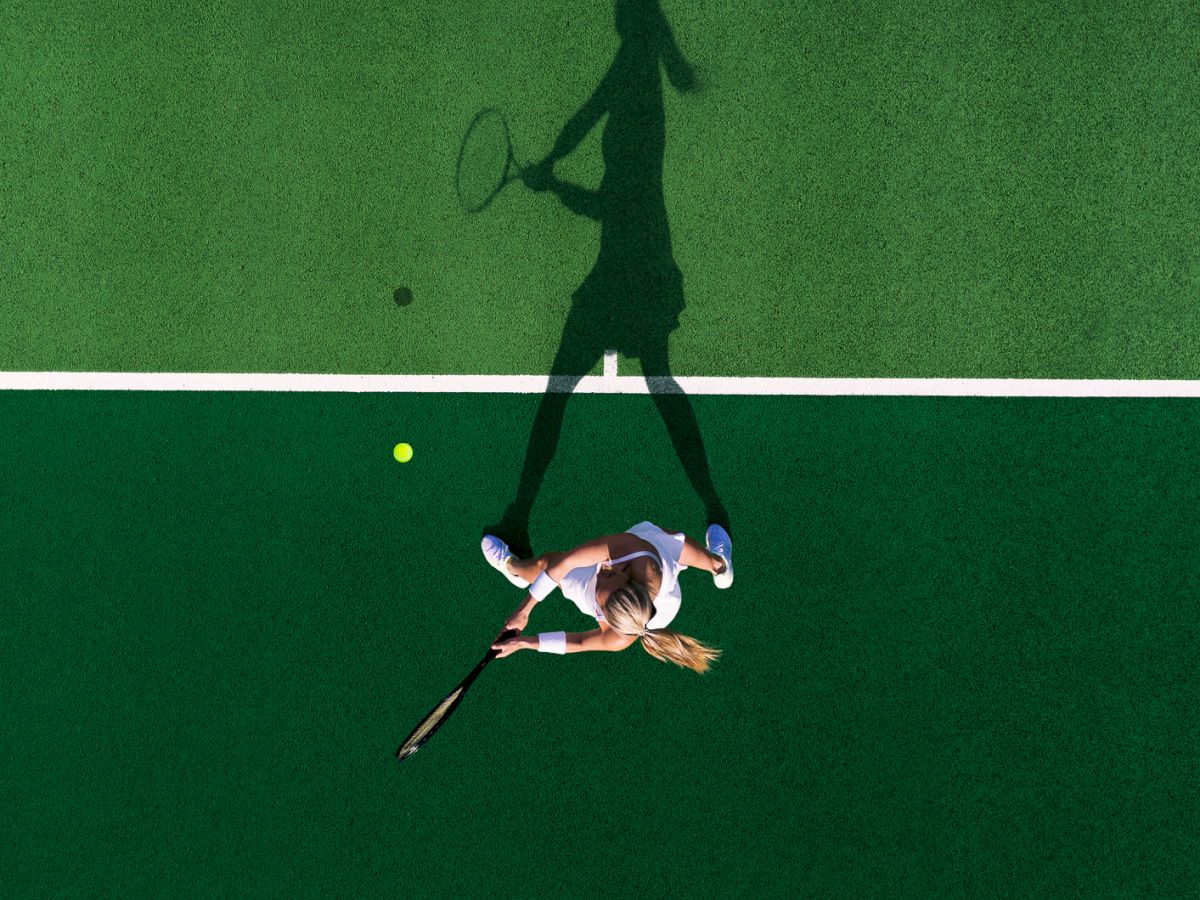 A person lies on a tennis court with a racket. Their shadow is poised to hit a ball, creating an artistic contrast.