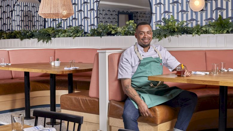 A person sitting on a booth in a restaurant with patterned walls and hanging lights, wearing an apron and holding a drink, smiling at the camera.