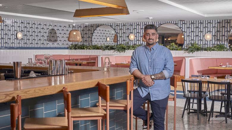 A person stands in a modern, stylish restaurant with wooden accents and patterned walls, featuring a spacious bar area.