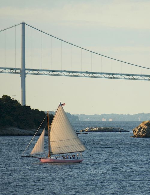 A sailboat on the water, with a house on a small island and a large bridge in the background, during daylight.