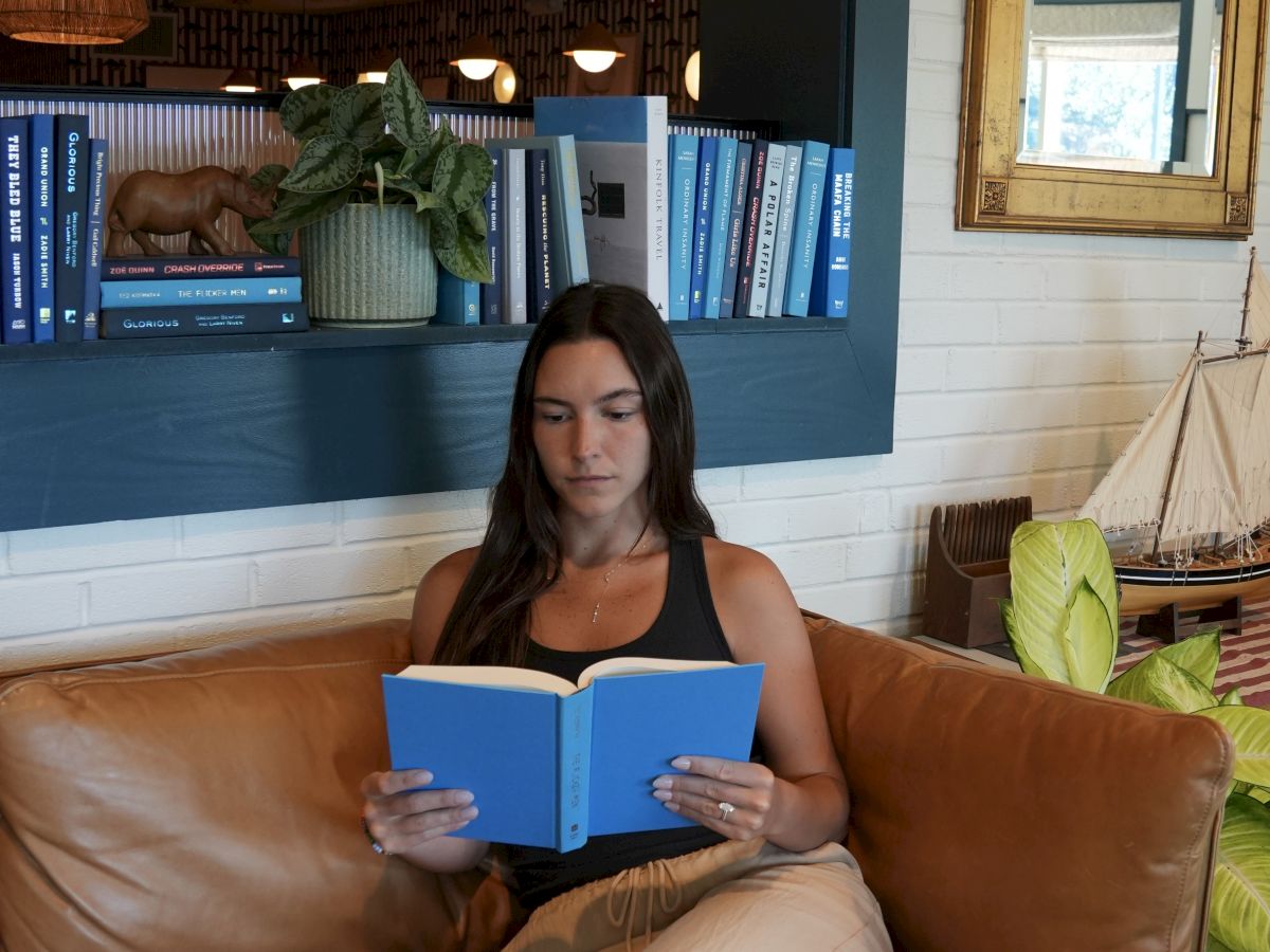 A person is sitting on a brown sofa reading a blue book in a cozy room with bookshelves and plants in the background.