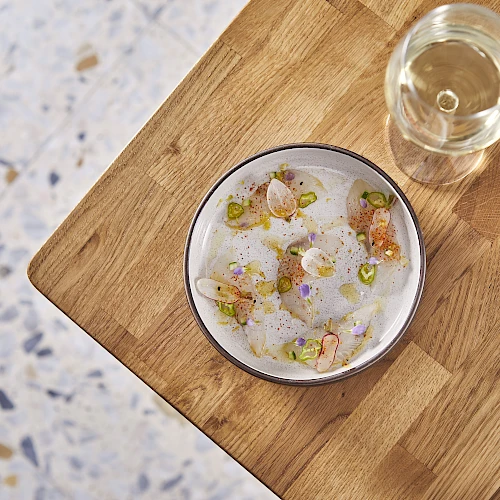 A small bowl with a delicate, plated dish next to a glass of white wine on a wooden table, bright and minimalist.