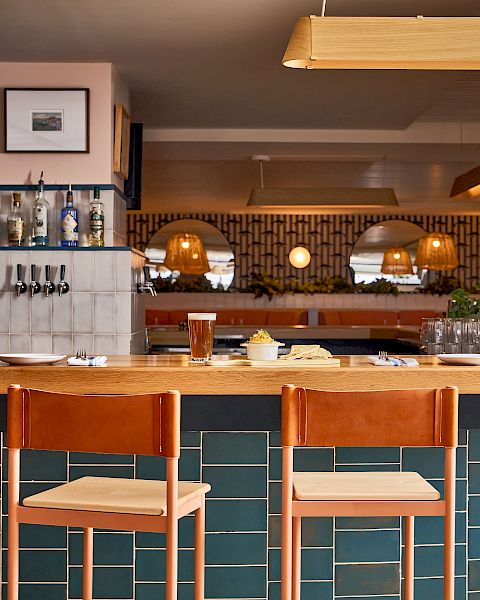 A stylish bar with a teal tiled counter, wooden stools, warm lighting, shelves of bottles, and a cozy seating area in the back.