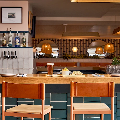 A stylish bar with a teal tiled counter, wooden stools, warm lighting, shelves of bottles, and a cozy seating area in the back.