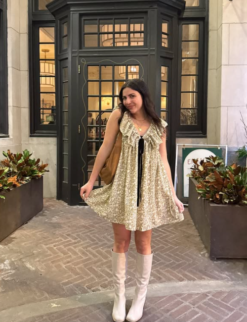 A person in a patterned dress and boots stands outside a building with plants and decorative doors, posing for the camera.