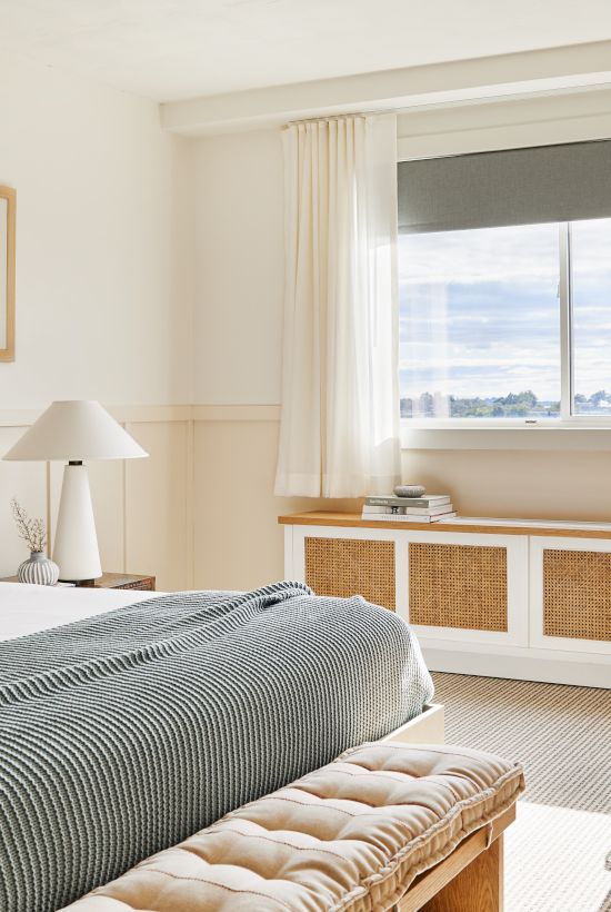 A cozy bedroom with a bed, chair, table, and large window, featuring warm lighting and minimalist decor, creating a relaxing atmosphere.