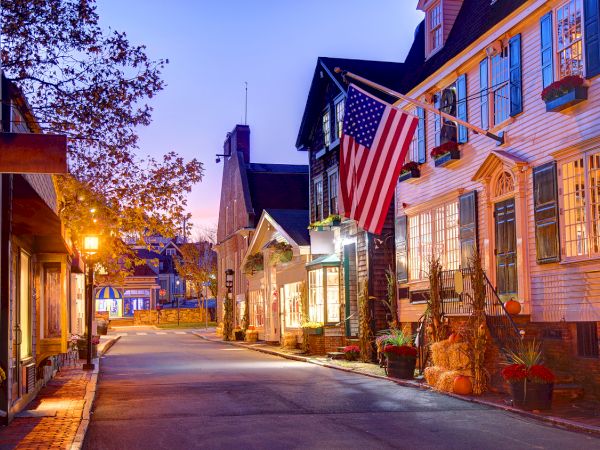 A quaint evening street scene features charming storefronts, an American flag, autumn decorations, and warm street lighting.