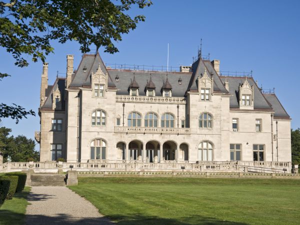 A large historic mansion with gothic architectural features, surrounded by a vast lawn and trees, set against a clear blue sky.