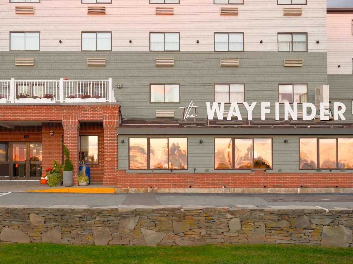 The image shows the exterior of a hotel named "The Wayfinder," featuring a brick and green facade, and large windows reflecting a sunset.