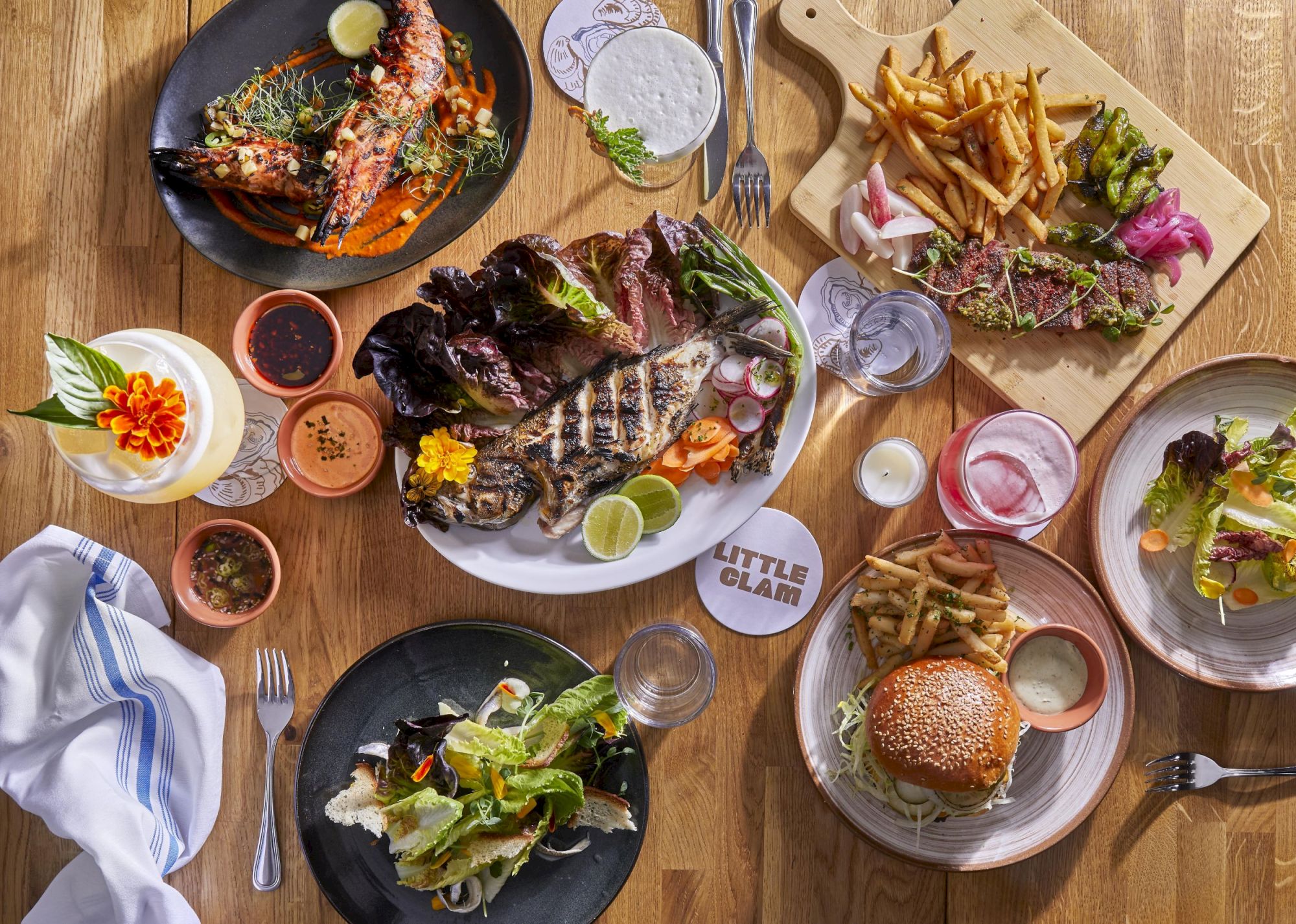 An assortment of dishes including burgers, salads, fries, steak, and drinks are displayed on a wooden table.