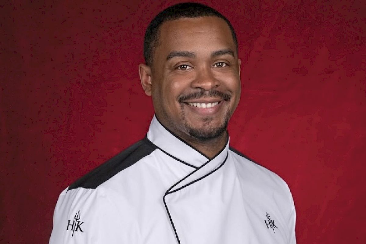 A person in a white chef's jacket with black accents is smiling against a red background, wearing a patch with "HK" on the sleeve.