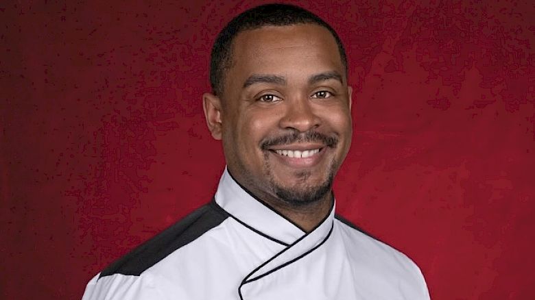 A person in a white chef's jacket with black accents is smiling against a red background, wearing a patch with "HK" on the sleeve.