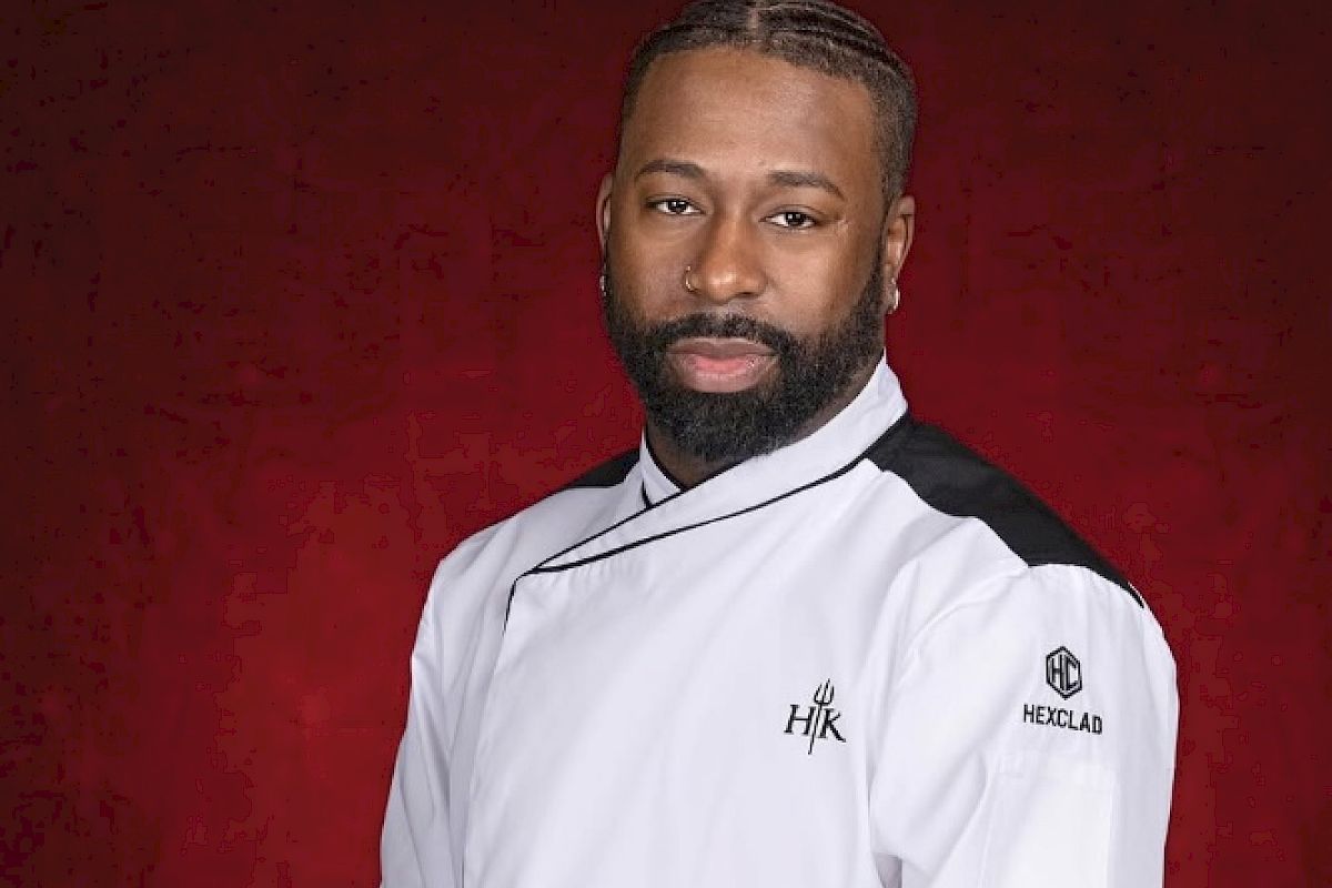 A person wearing a white chef's jacket with folded arms, standing against a red background.