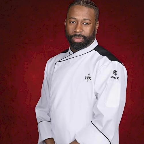 A person in a chef's uniform stands against a red background, looking at the camera with arms crossed in front of them.