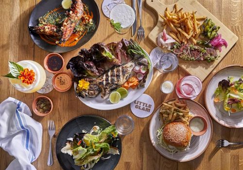 An array of dishes on a wooden table, including grilled meat, salad, a burger, fries, and drinks, creating a vibrant meal spread.