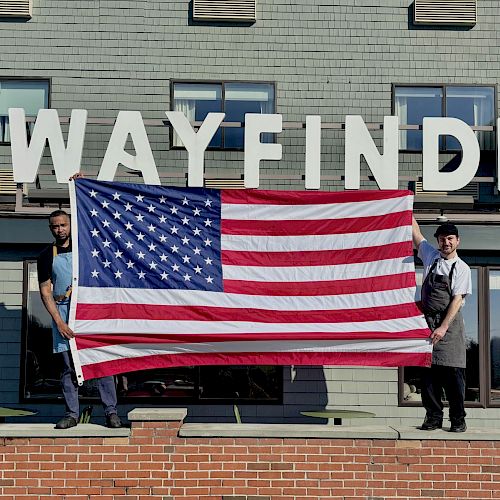 Two people are holding a large American flag in front of a building with "The Wayfinder" sign.