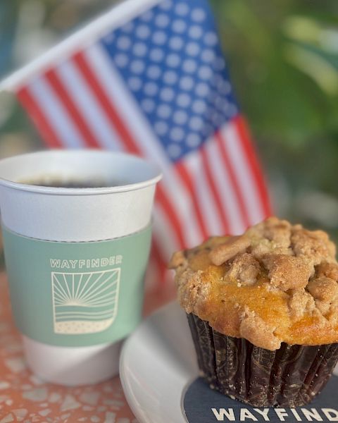 A coffee cup and muffin on a plate, with an American flag in the background, sit on a speckled tabletop.