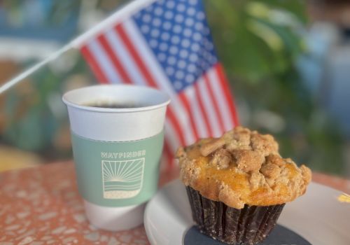 A coffee cup and muffin on a plate, with an American flag in the background, sit on a speckled tabletop.