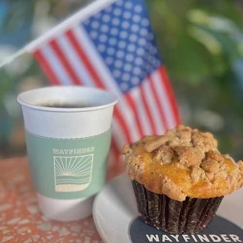A coffee cup and muffin on a plate, with an American flag in the background, sit on a speckled tabletop.