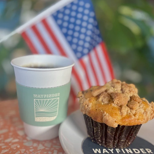 A coffee cup and muffin on a plate, with an American flag in the background, sit on a speckled tabletop.