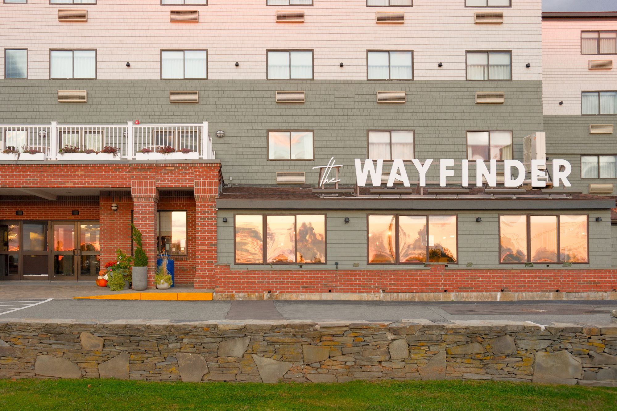 The image shows the exterior of a hotel building called "The Wayfinder" with large windows and a brick and green facade.