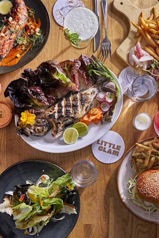 An assortment of dishes, including grilled meats, burgers, salad, and fries, are arranged on a wooden table for a meal.