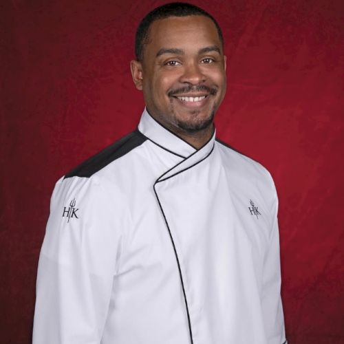 Guy in a chef's jacket with a red background, smiling at the camera.