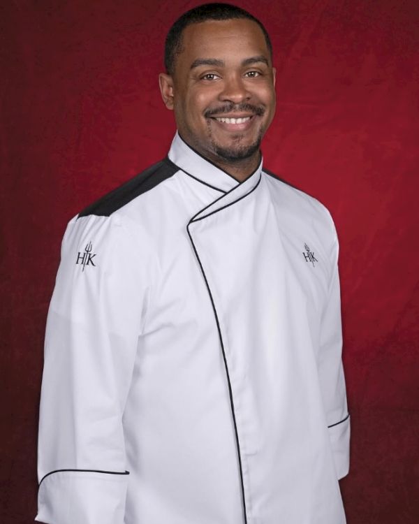Guy in a chef's jacket with a red background, smiling at the camera.