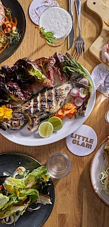 An overhead view of a table with various dishes, including seafood, salad, burger with fries, and drinks on a wooden surface.