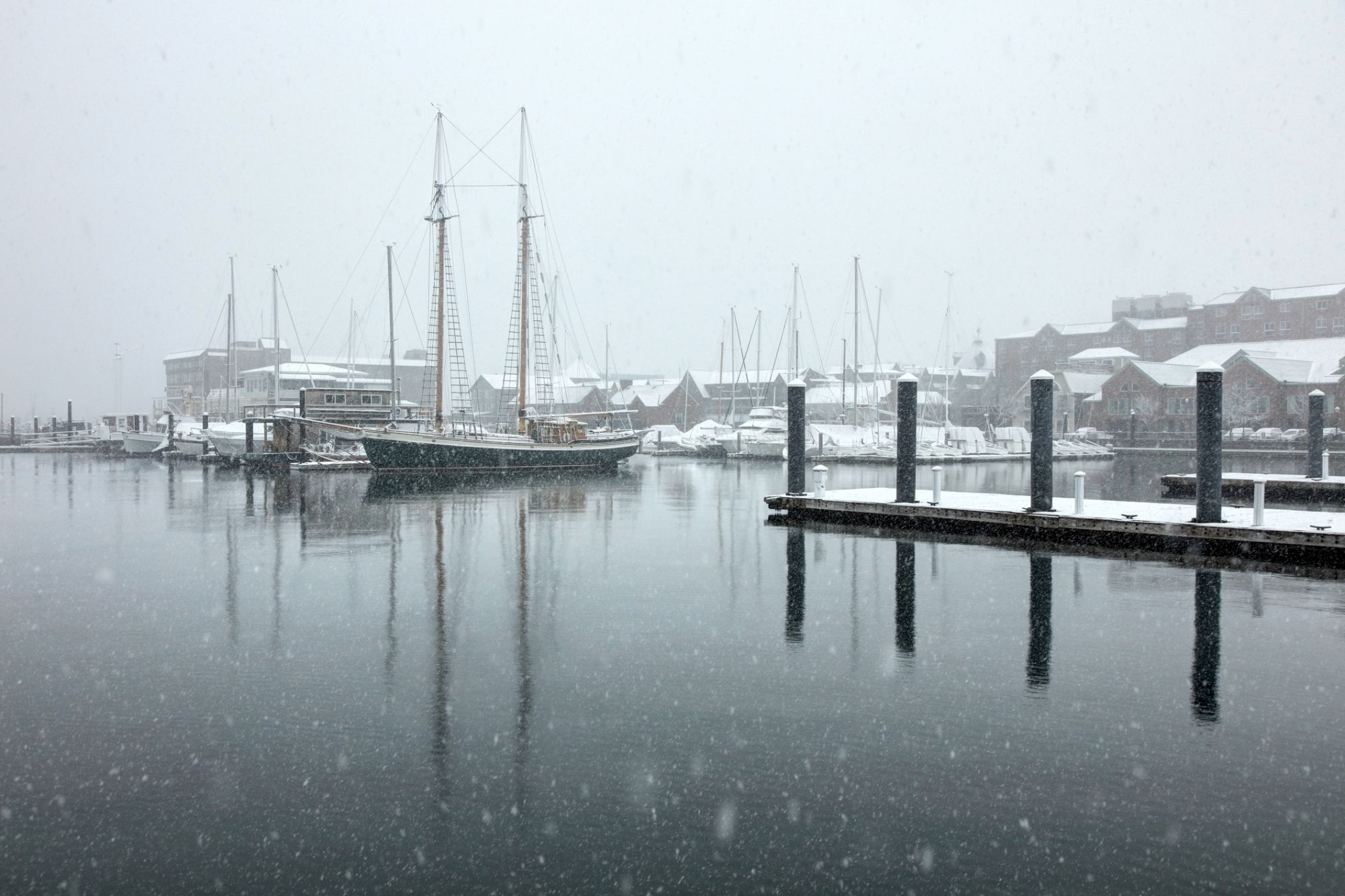 A snowy harbor scene with calm water, snowflakes falling, wooden docks, and a few sailboats and buildings fading into the mist.
