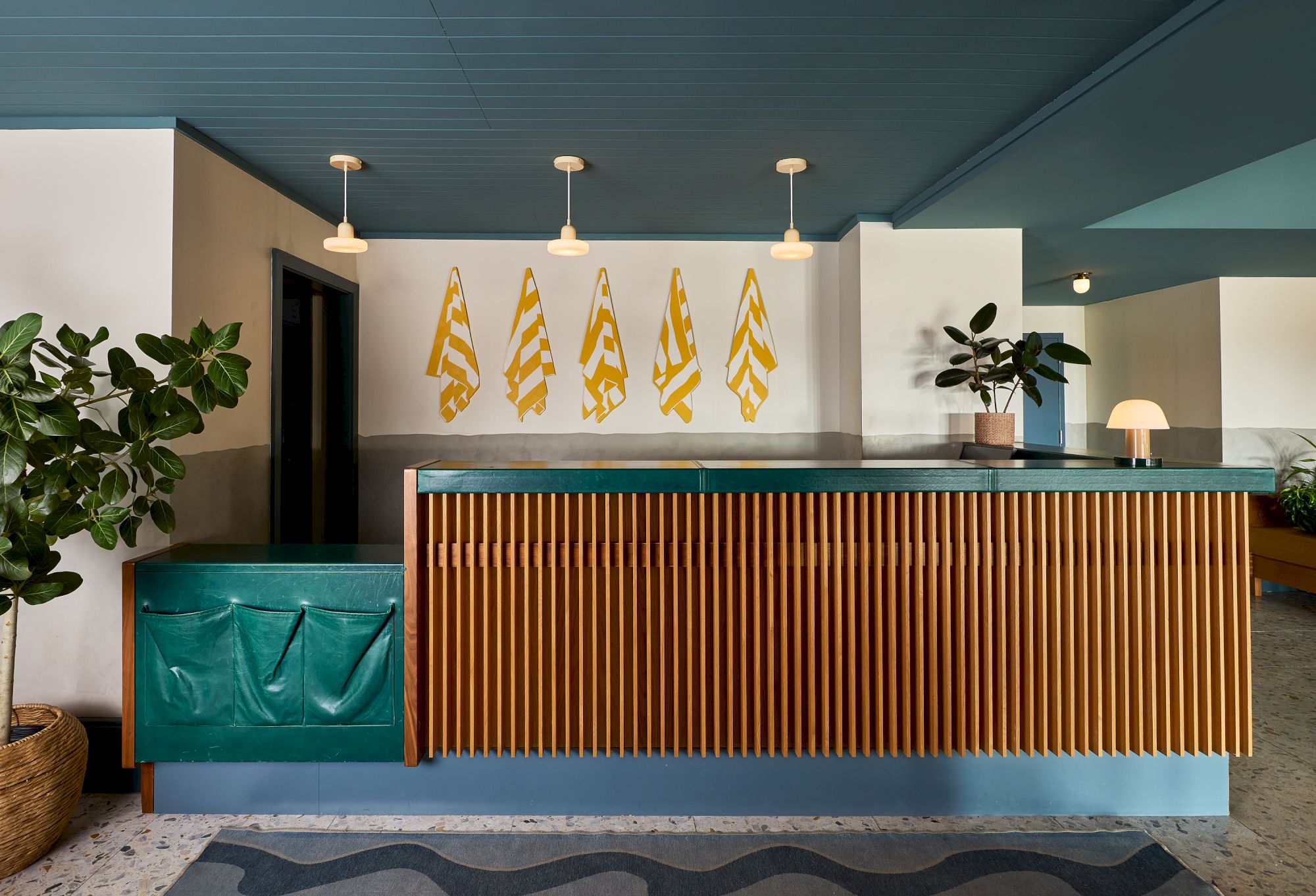 A modern hotel reception with a wooden slatted front, tropical plants, teal accents, and yellow zigzag wall art behind the desk, bright lighting.