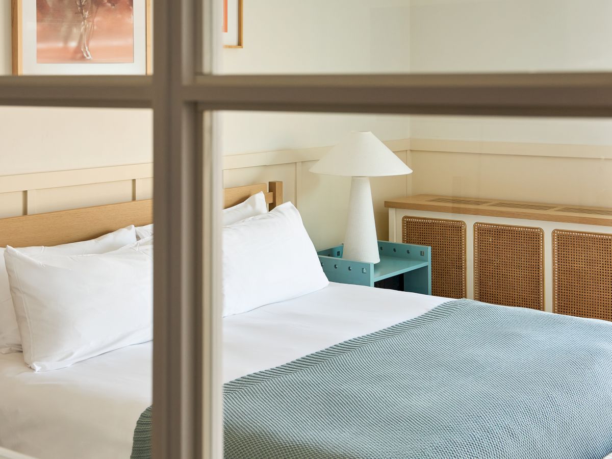 A cozy hotel room with a neatly made bed, white pillows, a light blue bedspread, a bedside lamp, and framed artwork on the wall, viewed through a window.