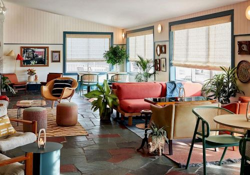 A cozy retro-style cafe or lounge with colorful seating, plants, and warm natural light streaming through blinds.