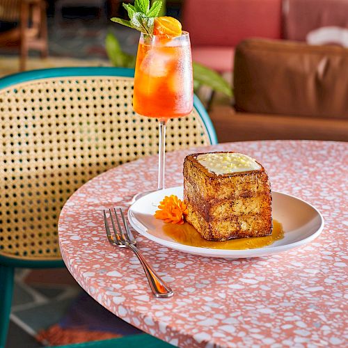 A glass of iced rose tea and a slice of layered cake on a plate, set on a pink terrazzo table with a colorful cafe backdrop.