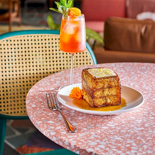 A glass of iced rose tea and a slice of layered cake on a plate, set on a pink terrazzo table with a colorful cafe backdrop.