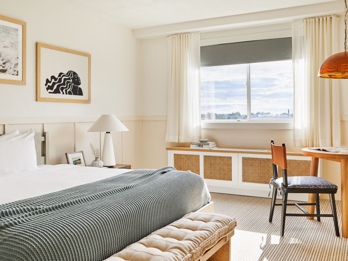 A bright hotel room featuring a bed with a striped comforter, a bench at the foot, a desk and chair by a window, and warm pendant lighting.