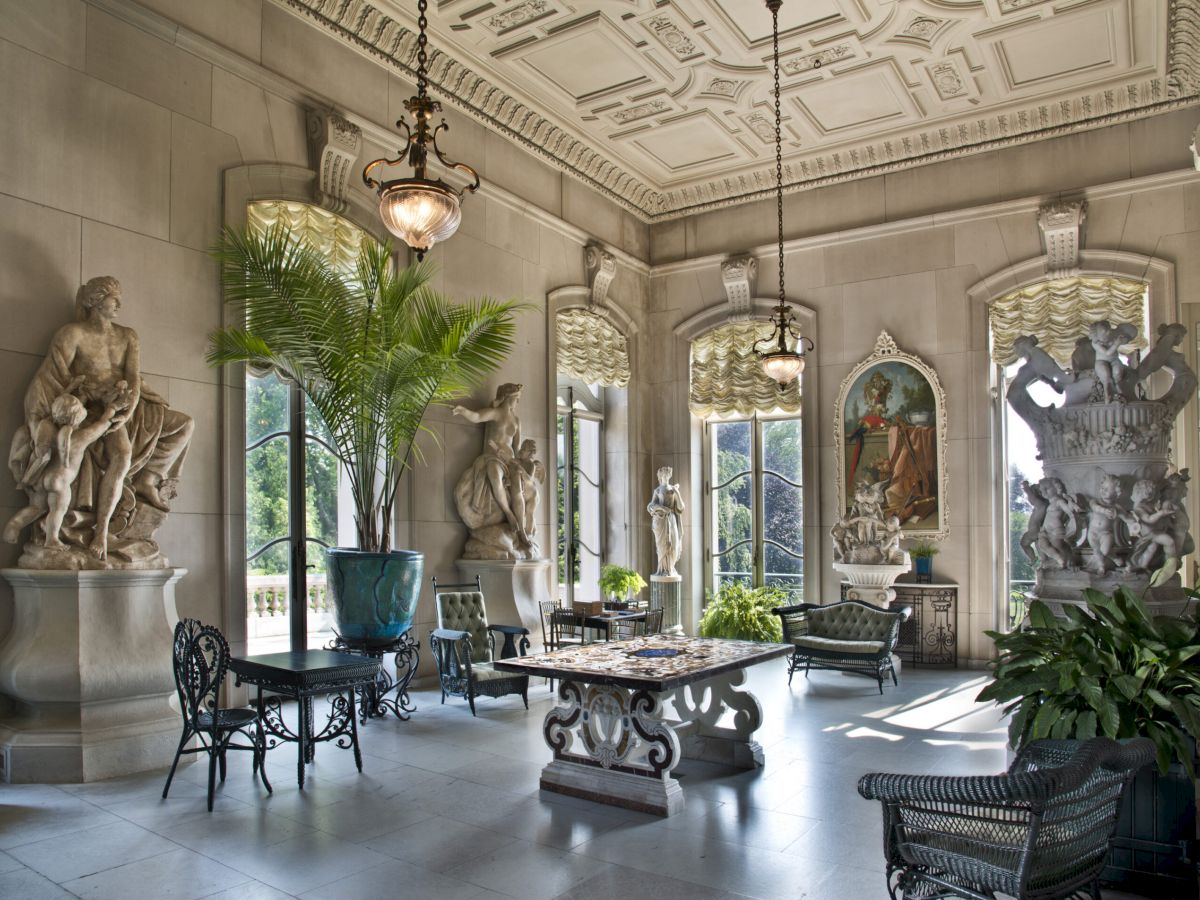 A grand, ornate interior with classical statues, arches, potted plants, and a central stone table in a sunlit, elegant space, ending with a period.