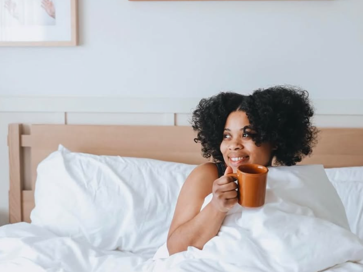 A woman sits up in bed, cozy under white sheets, sipping coffee from an orange mug, framed art on the wall behind her.