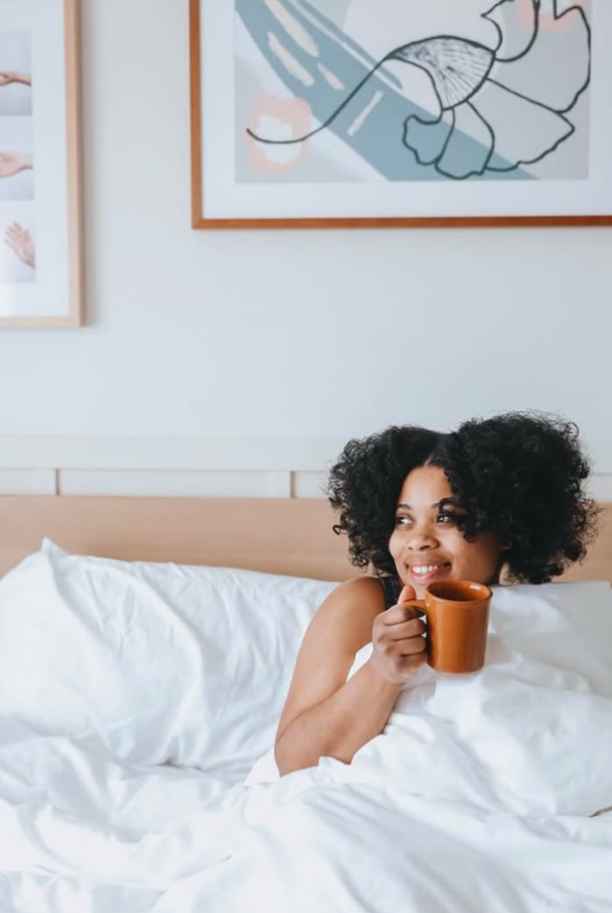 A woman sits up in bed, cozy under white sheets, sipping coffee from an orange mug, framed art on the wall behind her.