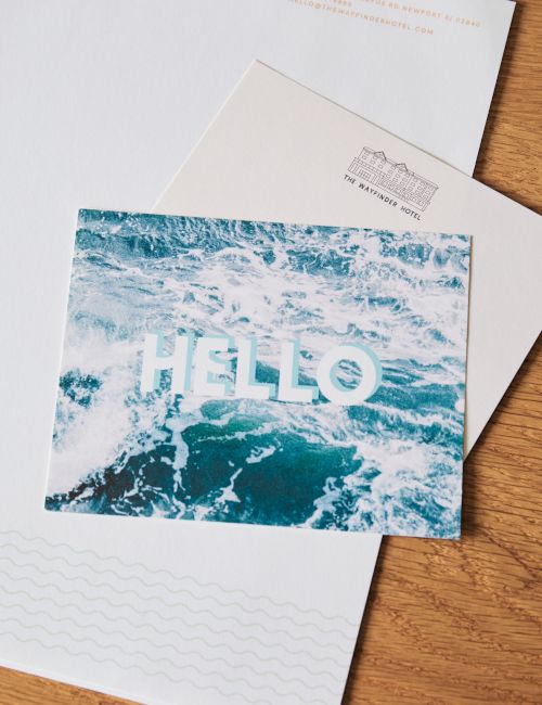 A postcard with a blue ocean wave image and the word &ldquo;HELLO&rdquo; on it, placed atop white envelopes on a wooden surface.
