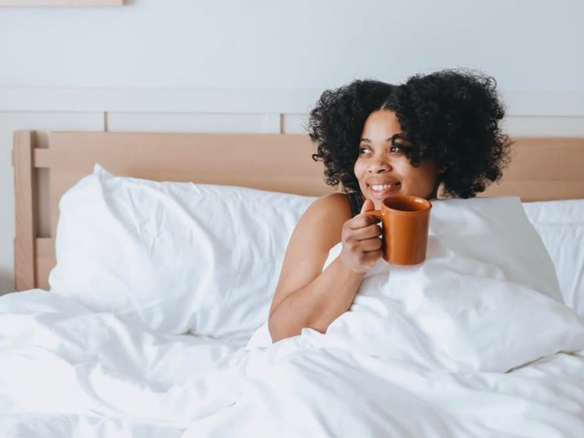 A smiling woman with curly hair sits up in bed, wrapped in white bedding, sipping from an orange mug, with framed art on the wall above.