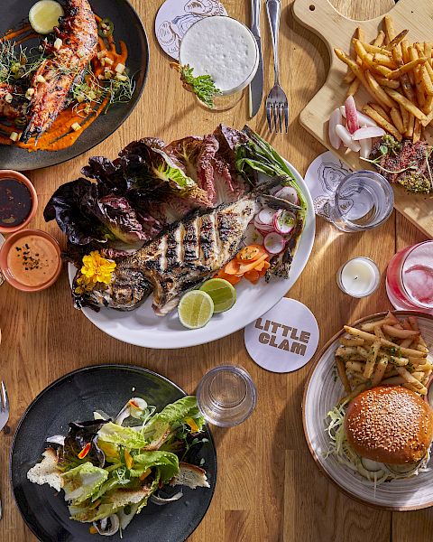 Sizzling plates of steak, burgers, fries, and fresh greens on a rustic wooden table&mdash;delicious spread for sharing.