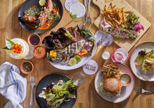 Sizzling plates of steak, burgers, fries, and fresh greens on a rustic wooden table&mdash;delicious spread for sharing.